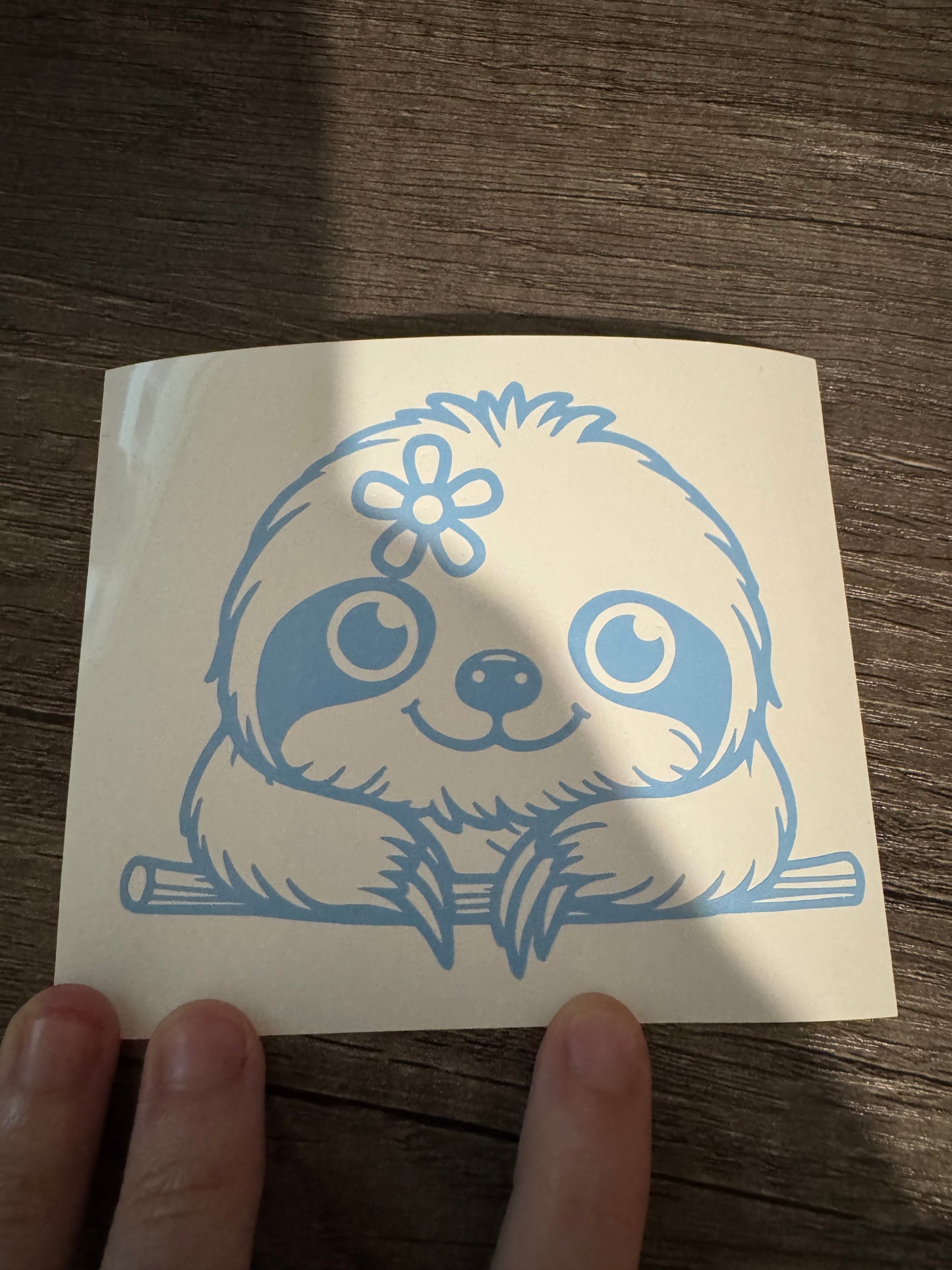 A blue decal of a smiling sloth with a flower on its head, held between fingers against a wooden background.
