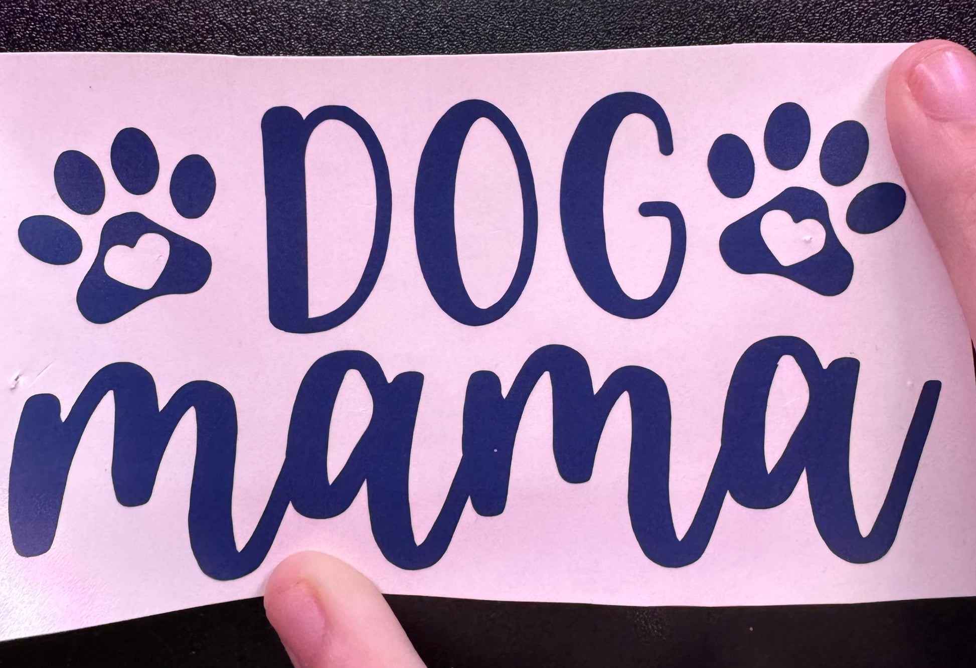 A custom vinyl decal with the text 'DOG mama' and paw prints, held between fingers on a white background.