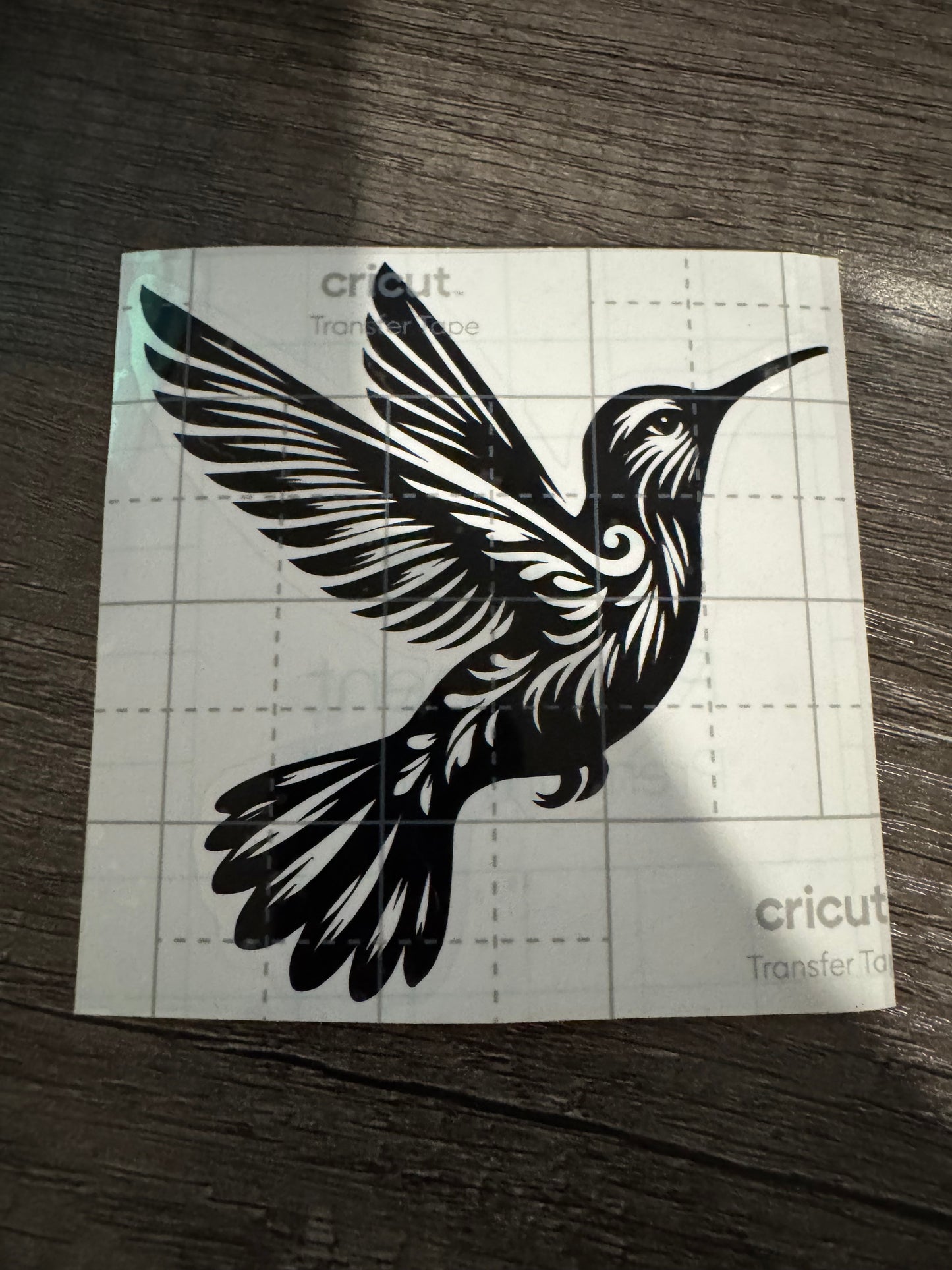 A black decal of a hummingbird placed on a white square surface with grid lines, likely a part of a crafting or customization process using a Cricut machine.