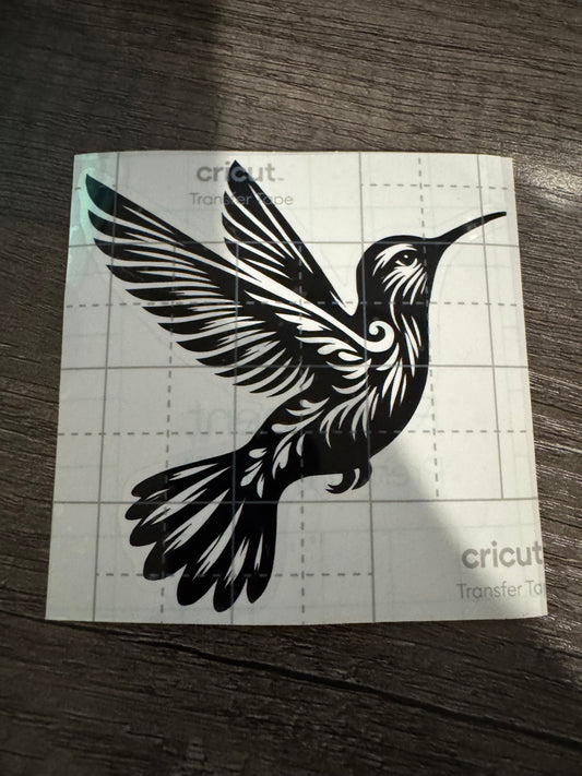 A black decal of a hummingbird placed on a white square surface with grid lines, likely a part of a crafting or customization process using a Cricut machine.