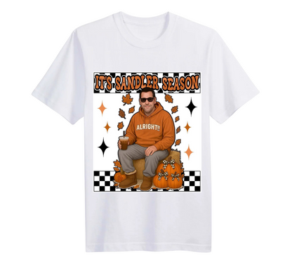 White t-shirt with a graphic of a man in an orange hoodie sitting on pumpkins and text 'It's Sandler Season' on a white background.